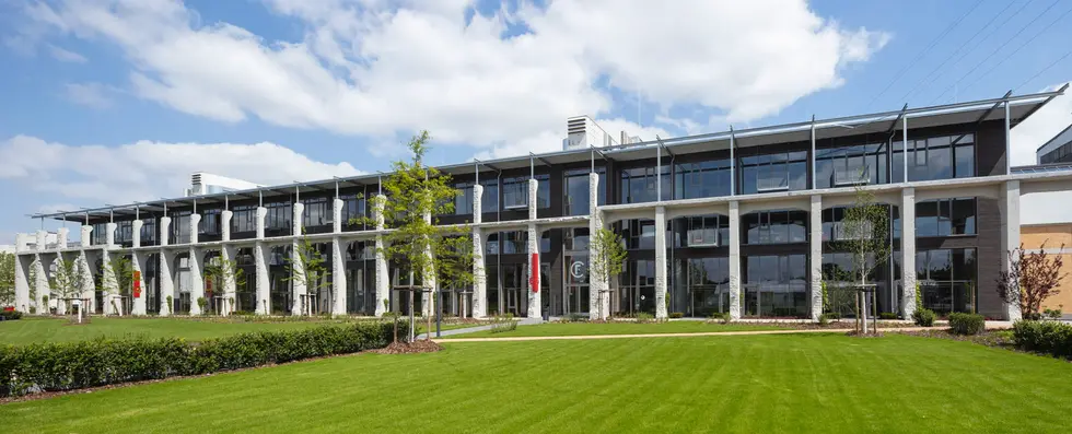 Arkadengebäude des Factory Campus Das Bild zeigt das Arkadengebäude des Factory Campus in Düsseldorf mit großen Fenstern, umgeben von gepflegtem Grün und Büschen. Der Himmel ist größtenteils blau mit einigen Wolken.