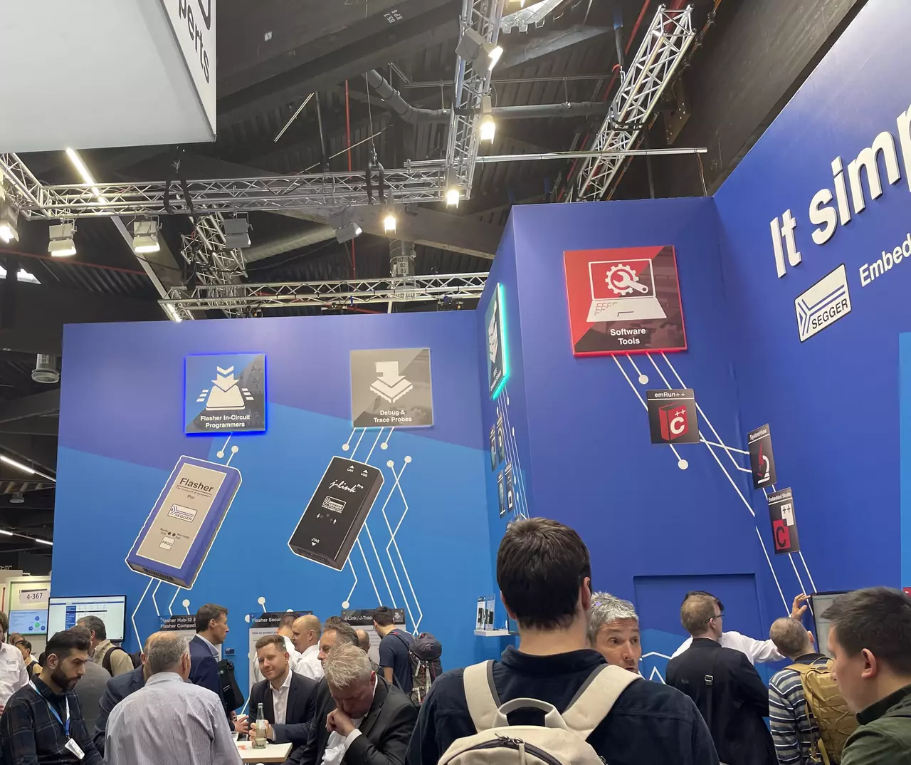 Das Bild zeigt einen Messestand, an dem mehrere Personen stehen und sich unterhalten. Im Hintergrund sind große, bunte Plakate mit Symbolen und Texten zu Software-Tools und Programmiergeräten an einer blauen Wand angebracht. Die Atmosphäre wirkt geschäftig und informell.