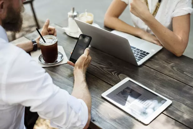 Zwei Menschen sitzen mit Smartphone, Tablet und Laptop gegenüber an einem Tisch