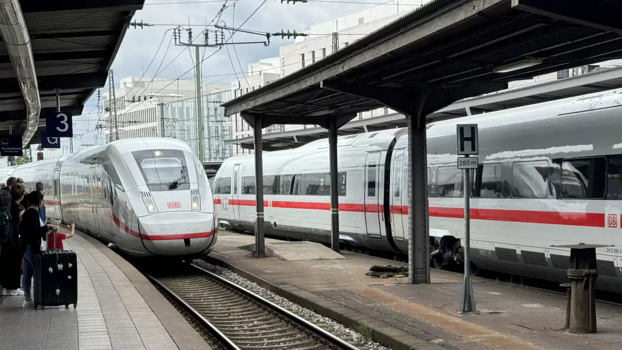 Ein moderner ICE-Zug der Deutschen Bahn fährt in einen Bahnhof ein. Am Bahnsteig stehen Reisende mit Gepäck. Im Hintergrund sind weitere ICE-Züge zu sehen, die auf dem Gleis stehen. Das Gleisbett ist sichtbar, umgeben von einem gepflasterten Gehweg.